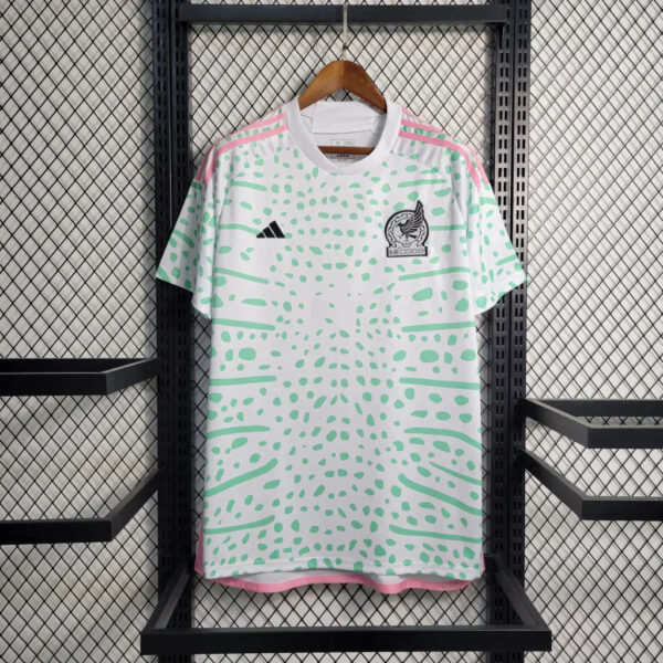 White Adidas Mexico soccer jersey with mint green abstract pattern and pink trim on a hanger against a metal grid background.