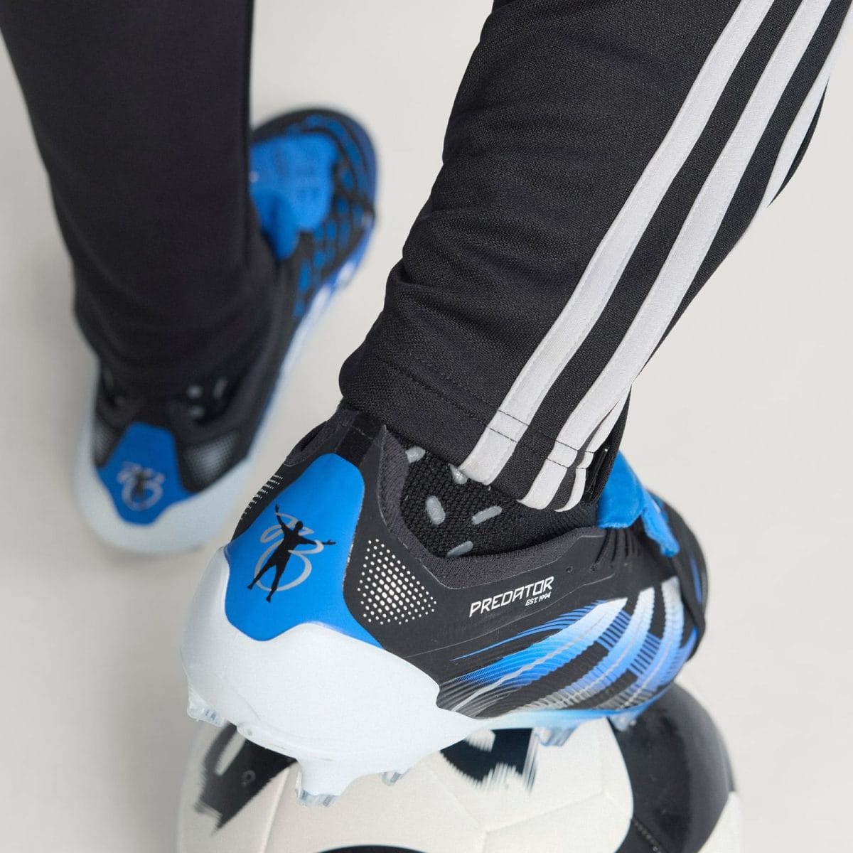 Close-up of a blue and black athletic shoe with 'Predator' branding, worn with black pants featuring white stripes.