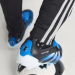 Close-up of a blue and black athletic shoe with 'Predator' branding, worn with black pants featuring white stripes.