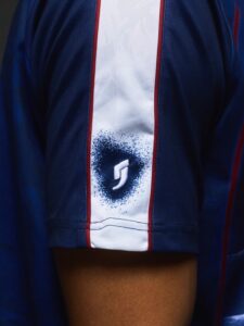 Close-up of a blue sports jersey sleeve with a white vertical stripe and a blue spray-paint circle logo containing a white stylized 'S'.