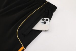 Black athletic shorts with gold piping; a white smartphone peeks out of the front pocket on a light surface.