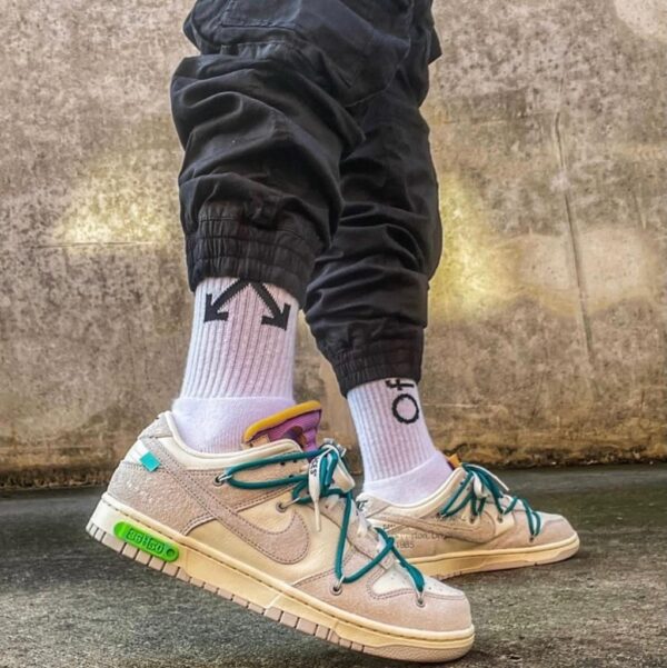 Person wearing tan sneakers with teal laces and white socks featuring arrows and 'OFF' branding, standing on a concrete surface in black joggers.