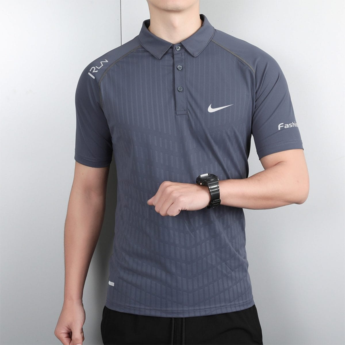 Person wearing a dark gray polo with Nike swoosh on the chest and a wristwatch on the left wrist, torso shot against a light wall.