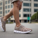 Close-up of a person wearing beige athletic running shoes stepping on pavement with an urban background behind them, mid-stride.
