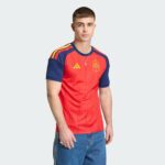 Young man in a Spain national team red jersey with blue sleeves, standing against a pale gray background.
