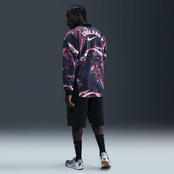 Person shown from behind wearing a dark patterned England soccer jacket, black shorts, black socks, and silver sneakers in a studio with a gray backdrop.