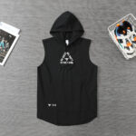 Black sleeveless hoodie with a front pocket and drawstring hood, laid flat on a gray surface with sneakers in a basket to the right and a magazine to the left; graphic text on chest reads, 'HARDEST WORKER IN THE ROOM.'
