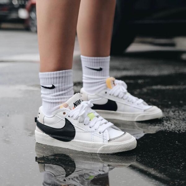 White Nike sneakers with black swoosh, worn with white socks, standing on a wet, reflective pavement.