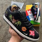 Black Nike sneaker with colorful embroidered patches (bats, starburst logos) held in hand; box with bold graphics in background.