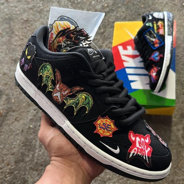 Black Nike sneaker with colorful embroidered patches (bats, starburst logos) held in hand; box with bold graphics in background.