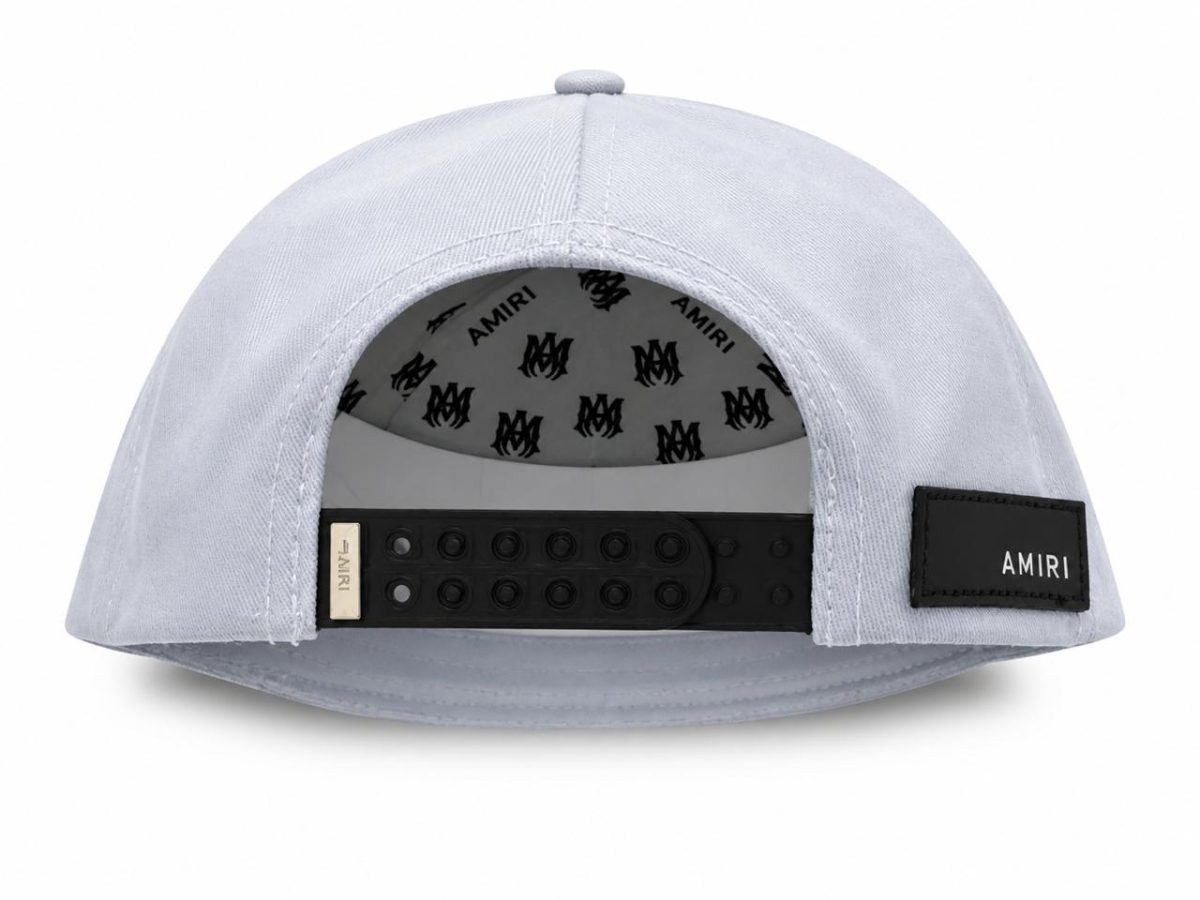 Light-gray snapback cap viewed from the back, showing a black plastic strap and a small Amiri label on a patch at the side edge of the opening.