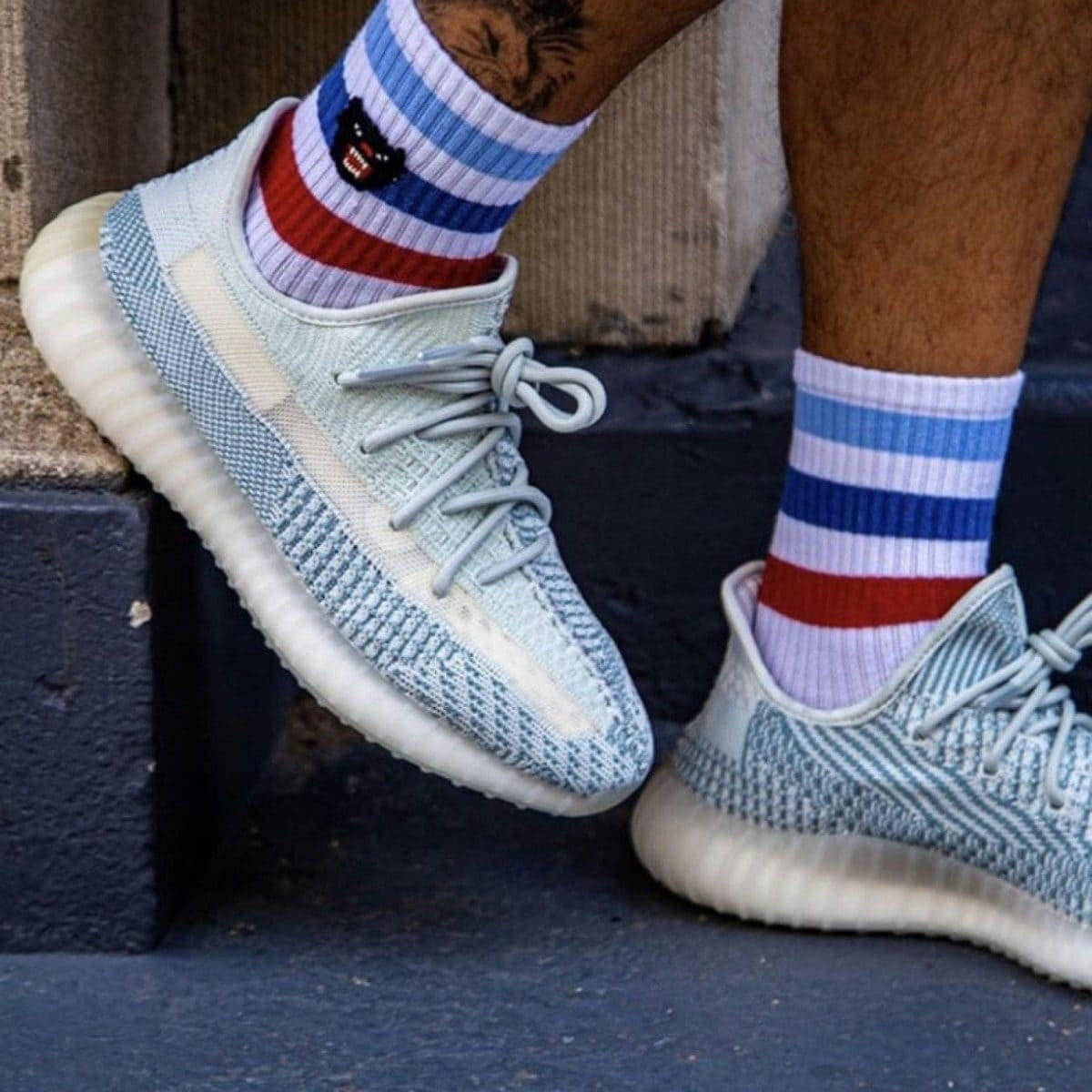 Light blue knit sneakers with pale soles, laced, worn with red, white, and blue striped crew socks on a dark step.