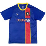 Blue soccer jersey with a red vertical stripe bordered in yellow, yellow-trim collar, crest on the chest, and the 'DUBAI VR' sponsor text across the front.