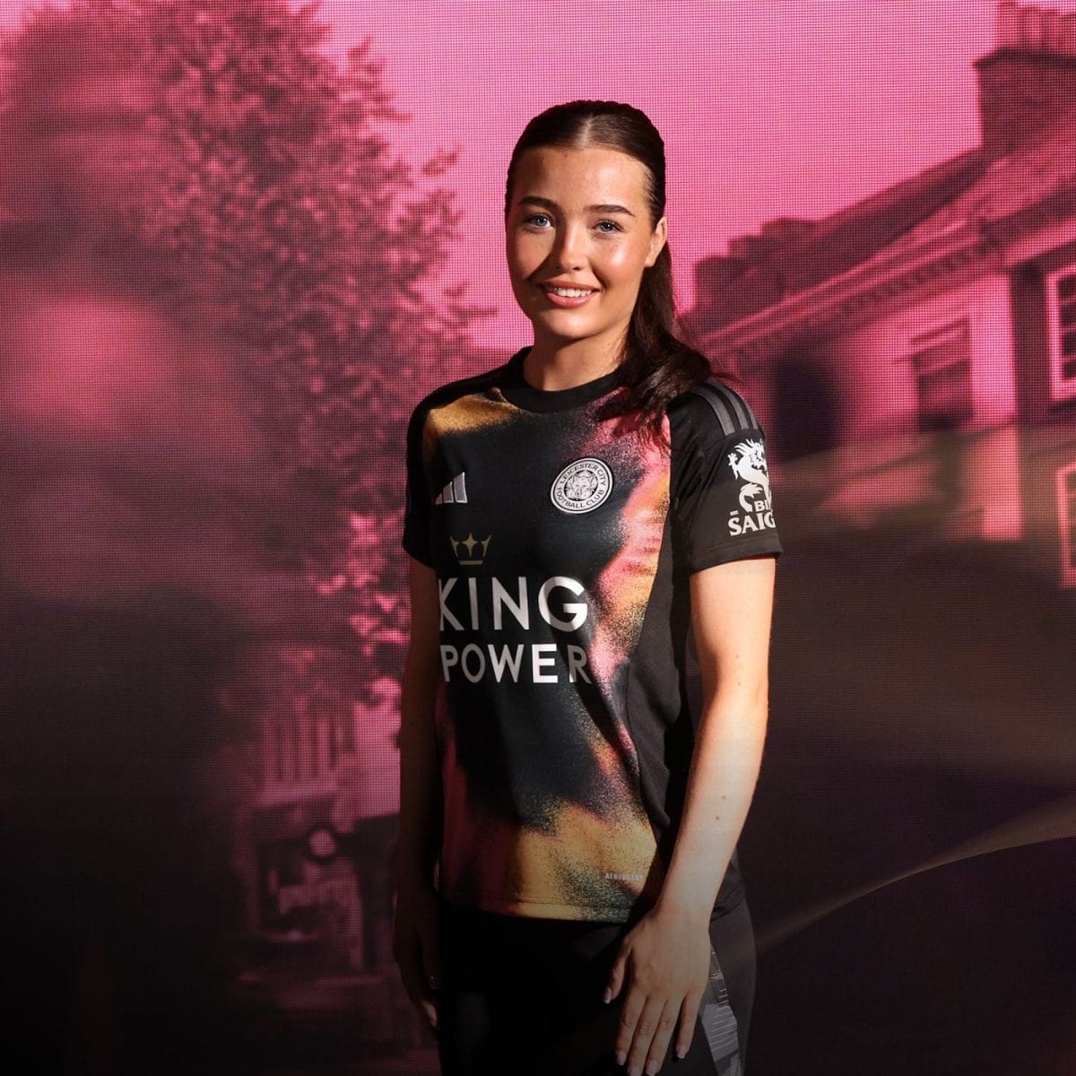 Smiling woman in a black Leicester City football jersey with a King Power logo, standing against a pink backdrop.