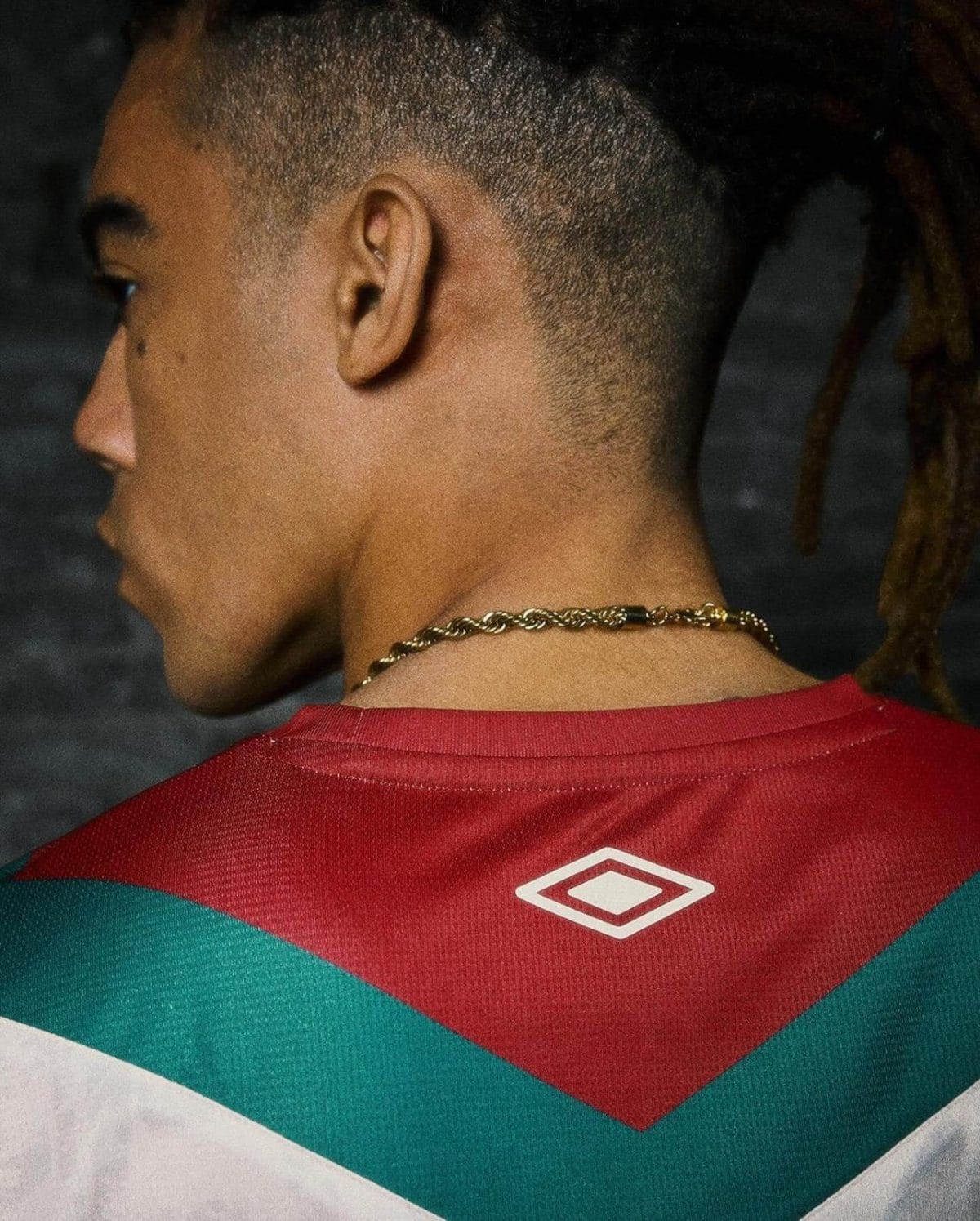 Profile of a person with a short fade and dreadlocks, wearing a red and teal geometric sports jersey and a gold chain necklace