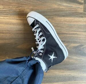 Black high-top canvas sneaker with white laces on a wooden floor, worn with blue pants; white toe cap and side star logo visible.