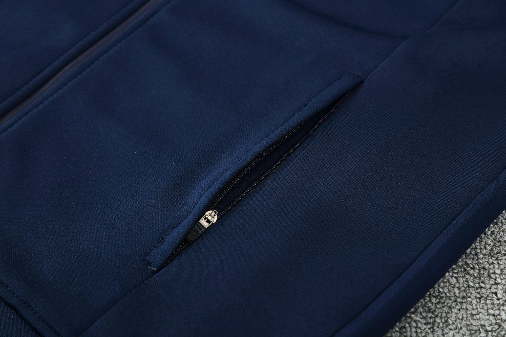 ba4c396b | UAE SPORT STORE Close-up of navy blue fabric with a silver zipper on a jacket laid on a gray carpet