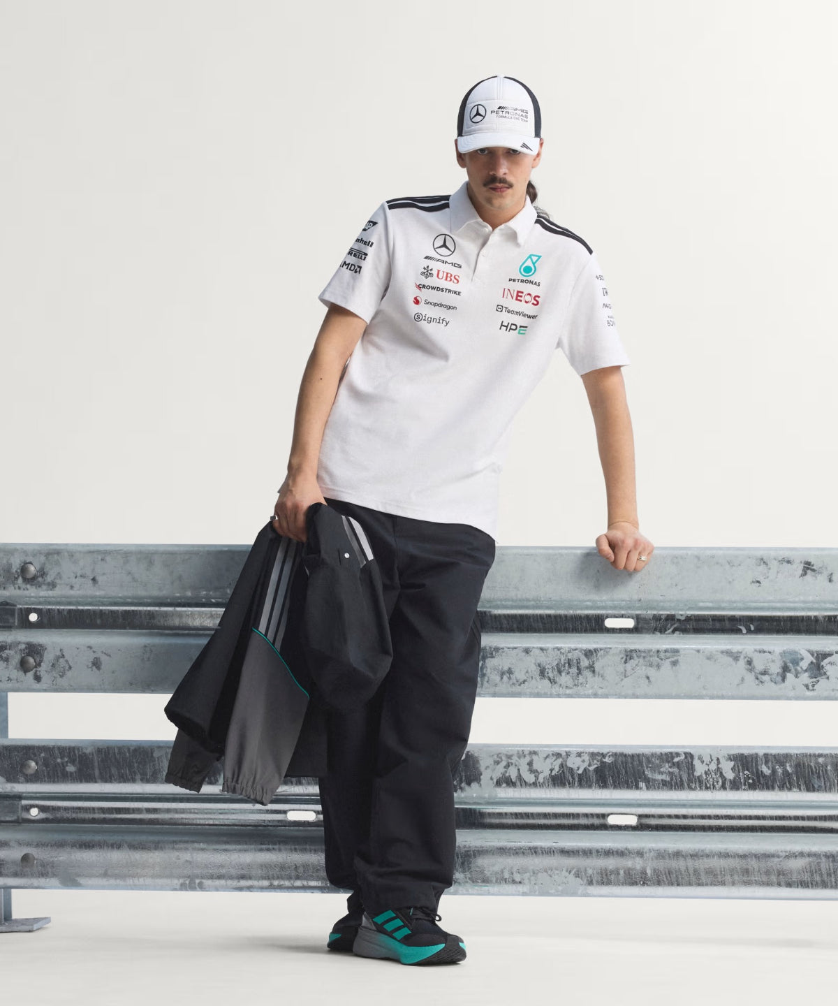 ba64e87504342e457c7667d8a59379f4-1200x1440-1 | UAE SPORT STORE Man leaning on a metal guardrail, wearing a white polo with logos, black pants, teal-accent sneakers, and a white cap, holding a jacket.