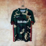 Camouflage FC Barcelona football jersey on a hanger, with Rakuten sponsor and Nike logo against a beige wall.