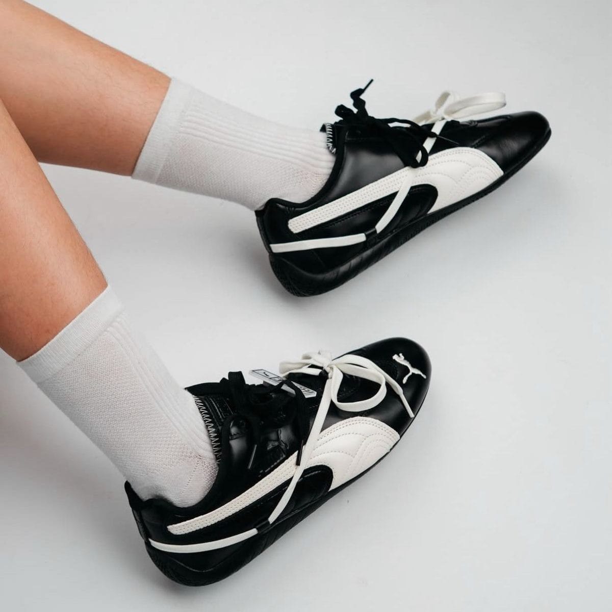 Pair of black and white sneakers with white laces and white socks on a light gray background, viewed from above, showing the shoes side by side.