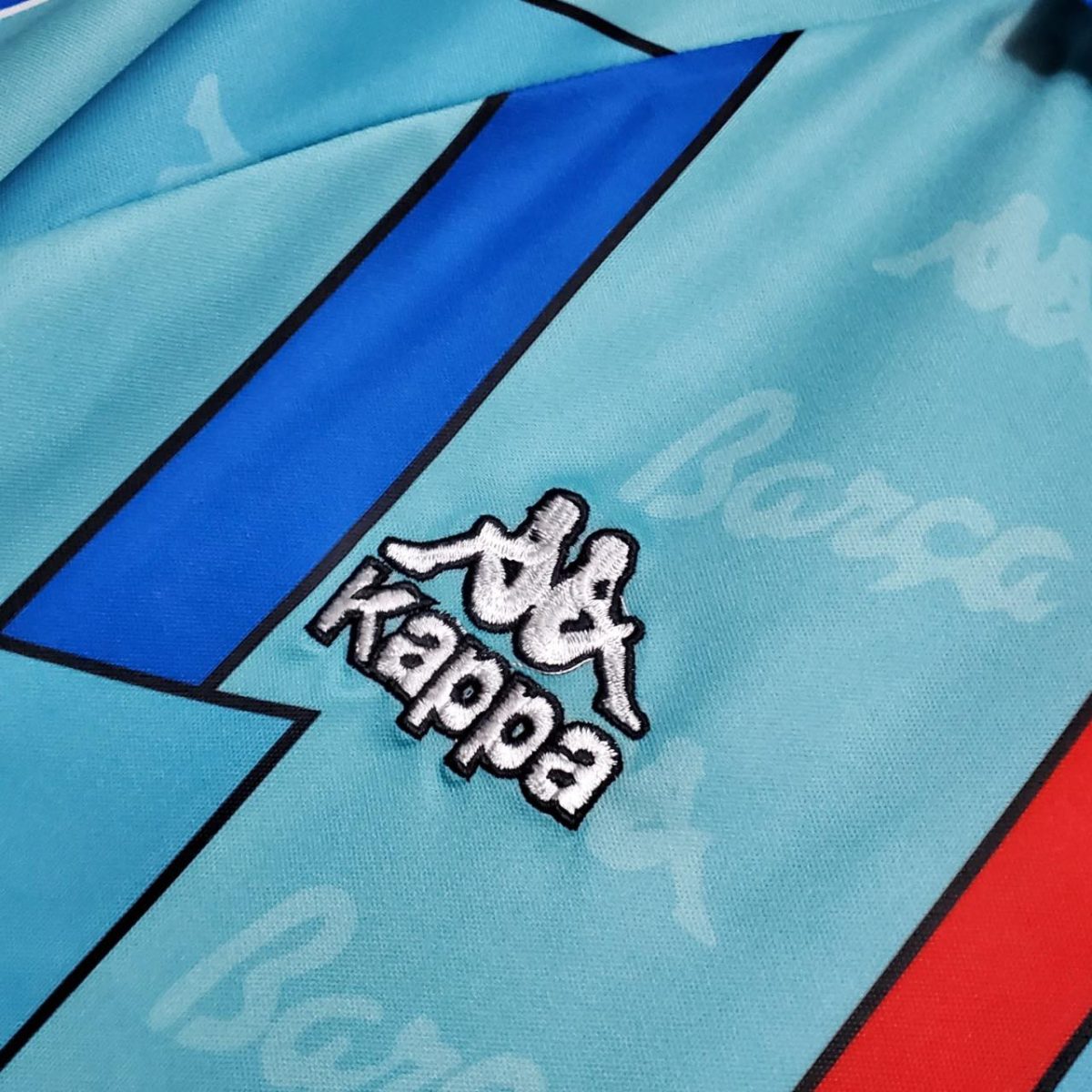 Close-up of turquoise fabric with a bold blue diagonal stripe and white embroidered Kappa logo.