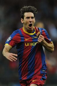Male soccer player in a blue and maroon Barcelona kit celebrates a goal, mouth open in a yell and fist clenched.
