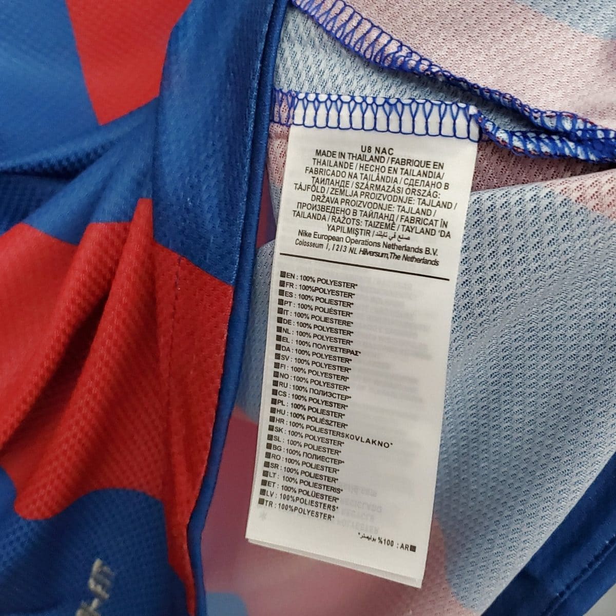 Close-up of a clothing tag on blue and red fabric, listing material and care instructions in multiple languages.