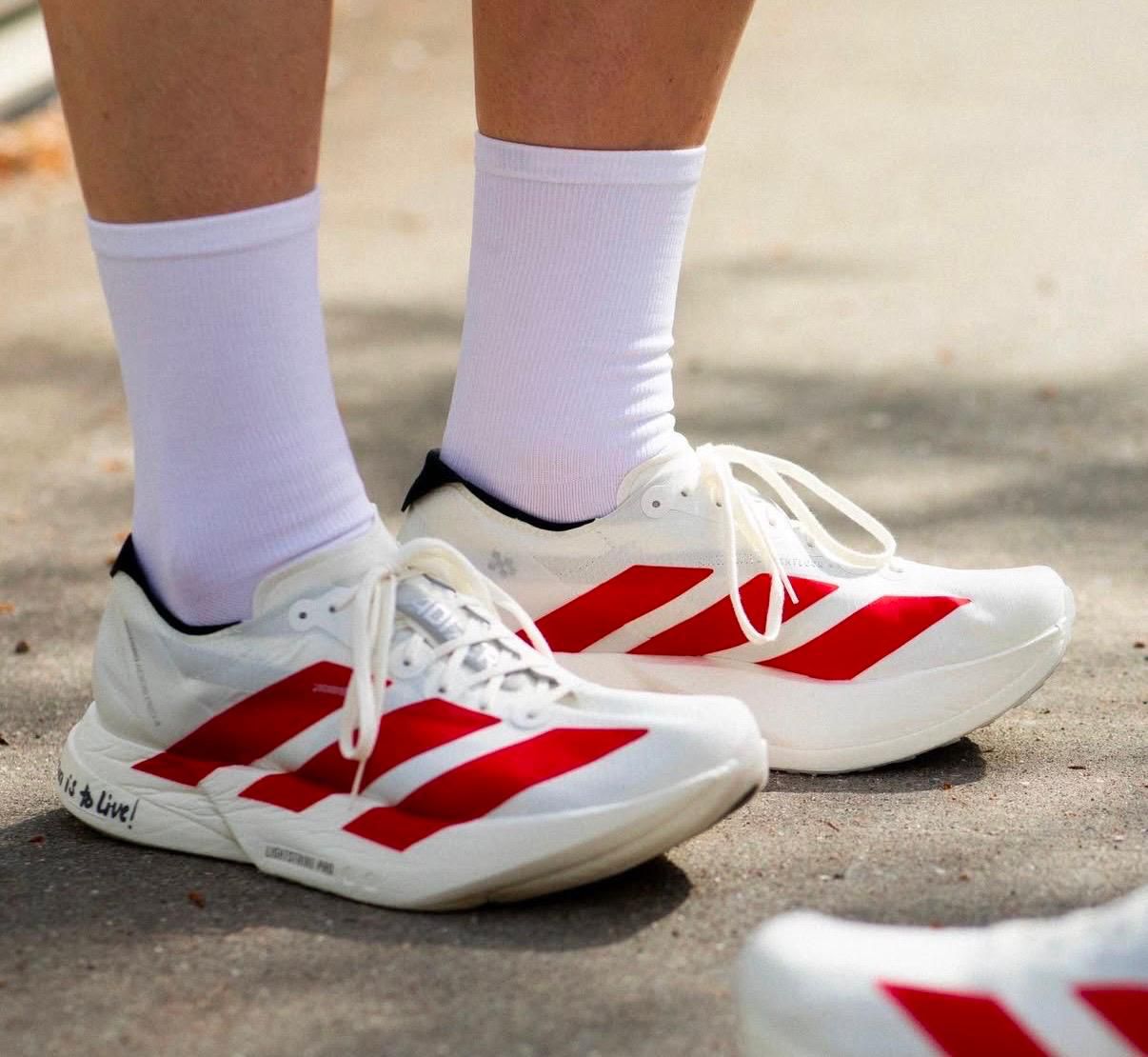bb2d6844-1778-4c7f-9307-638a32daa5ba Close-up of white athletic shoes with bold red stripes and white socks on a paved surface.