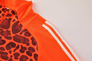 Close-up of a bright orange athletic jacket with a black leopard-print panel on the front and white double stripes along the sleeve near the shoulder.