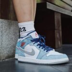Side view of white and light-blue Air Jordan 1 sneakers with blue laces, paired with a white sock showing '23'.