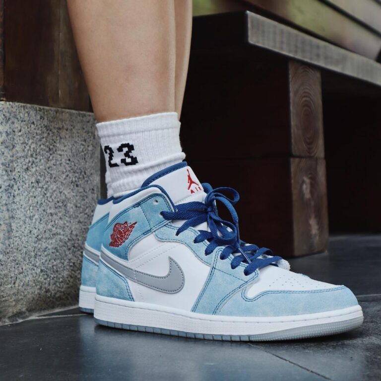 Side view of white and light-blue Air Jordan 1 sneakers with blue laces, paired with a white sock showing '23'.