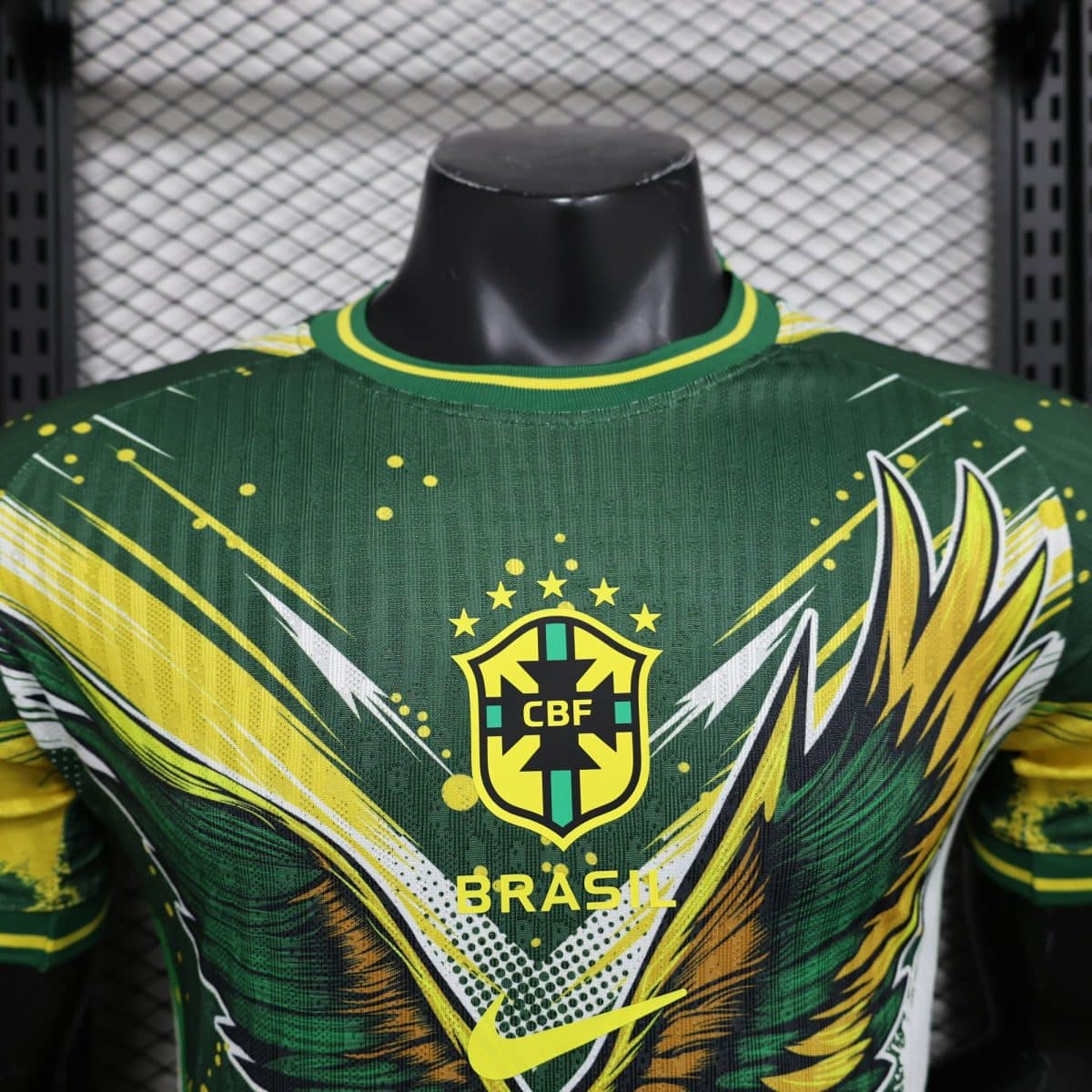 Close-up of a Brazil football jersey on a mannequin, green with yellow accents and the CBF crest, against a chain-link fence backdrop.