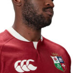 Close-up of a man wearing a red rugby jersey with a crest on the chest, showing part of his beard and neck.