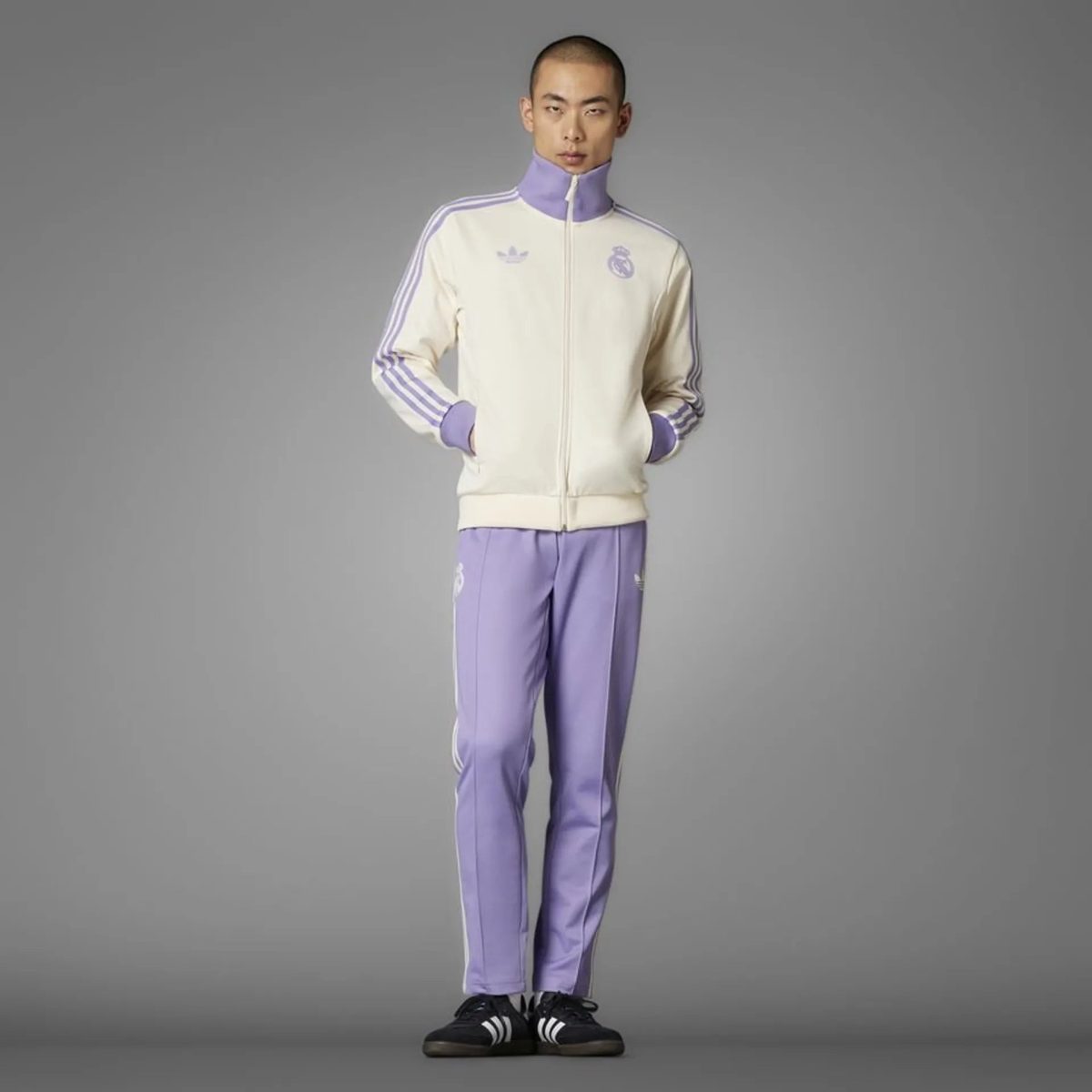 Full-body shot of a man wearing a cream track jacket with purple accents and matching lavender pants, standing with hands in pockets.