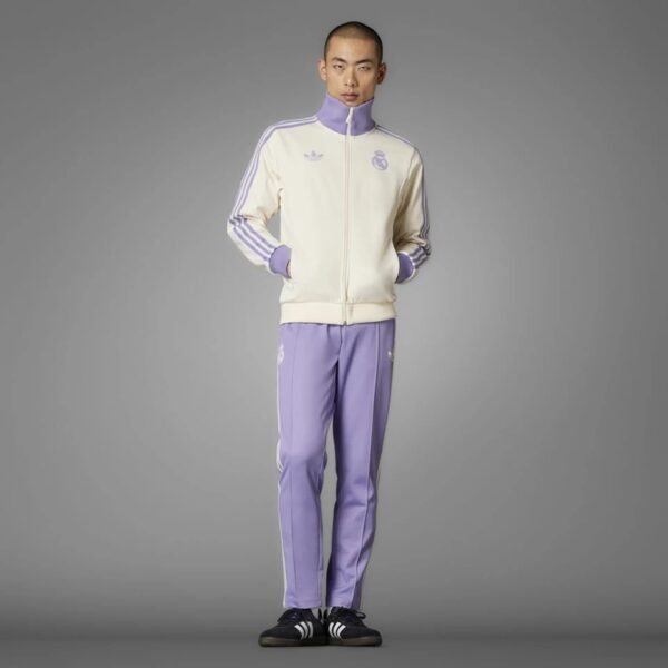 Full-body shot of a man wearing a cream track jacket with purple accents and matching lavender pants, standing with hands in pockets.