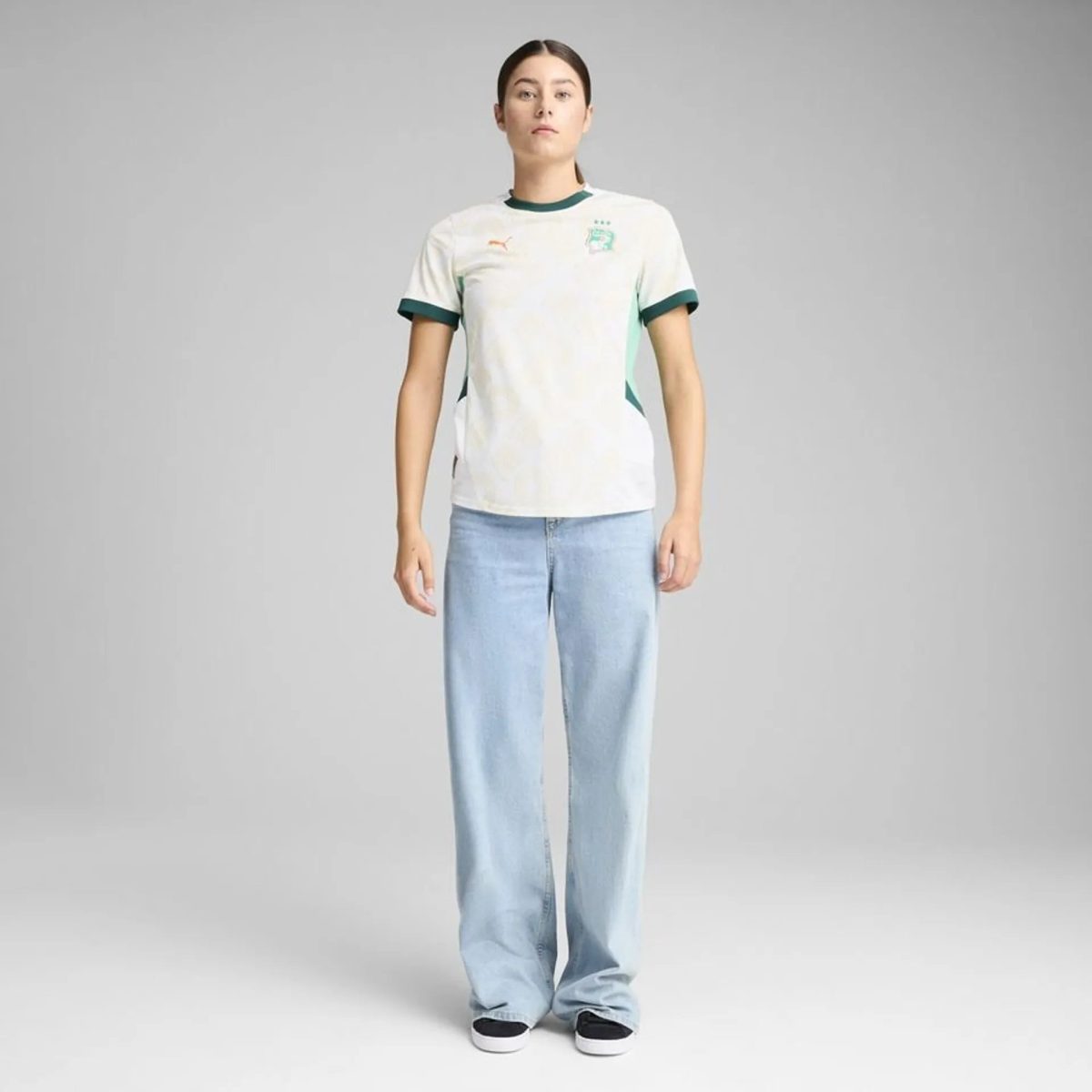 Full-body shot of a woman standing against a gray backdrop in a white jersey with green trim and light blue jeans, white sneakers.