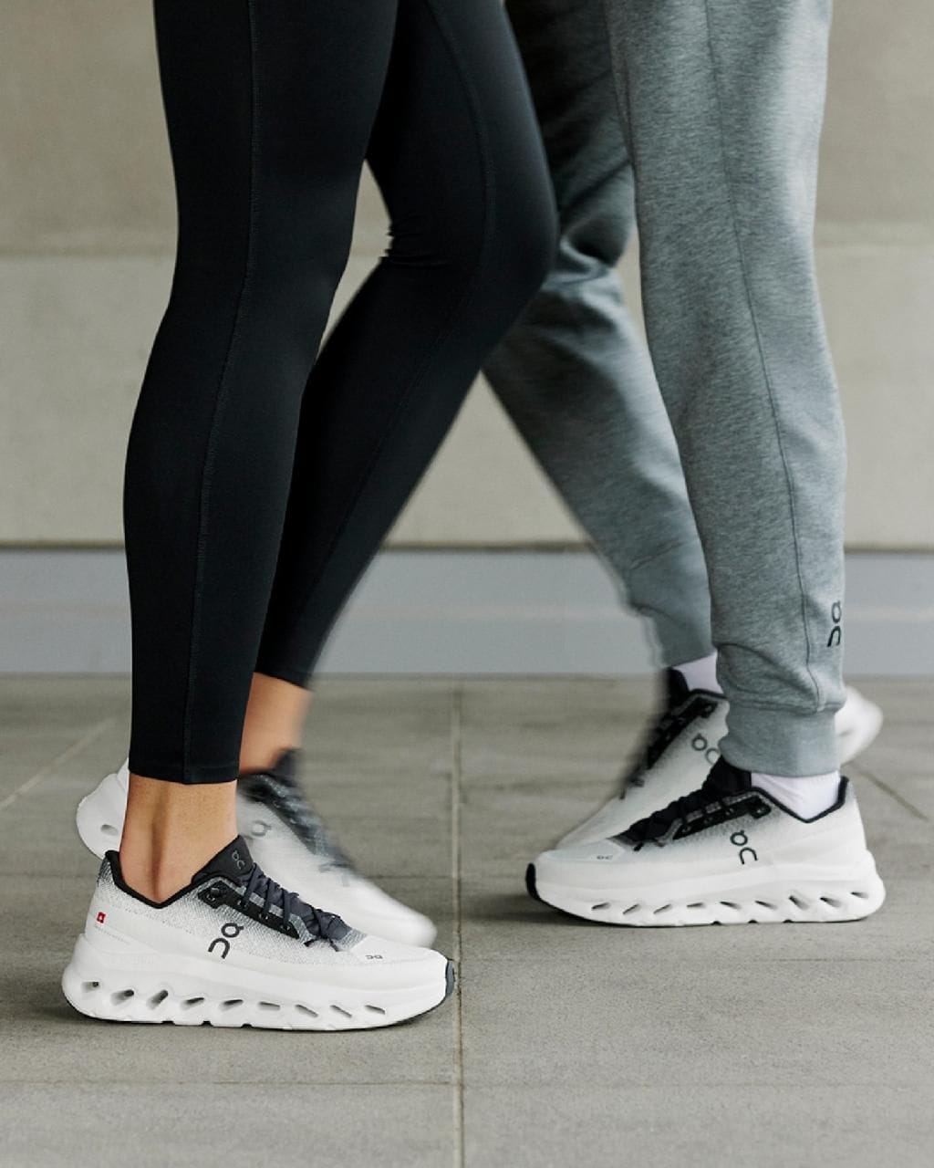 bd106fce-1d6a-47b6-87ec-d95d8991da6e | UAE SPORT STORE Two people wearing white athletic sneakers with chunky hollow soles, one in black leggings and the other in grey joggers, standing on a tiled surface.