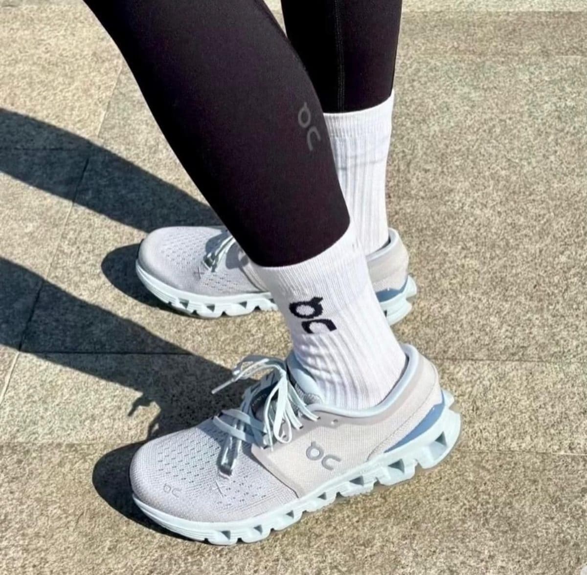 Person wearing white On running shoes and white socks with the On logo, standing on a concrete pavement in black leggings.