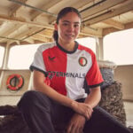Young woman in a red-and-white soccer jersey sitting on a rope on a boat deck, smiling at the camera.