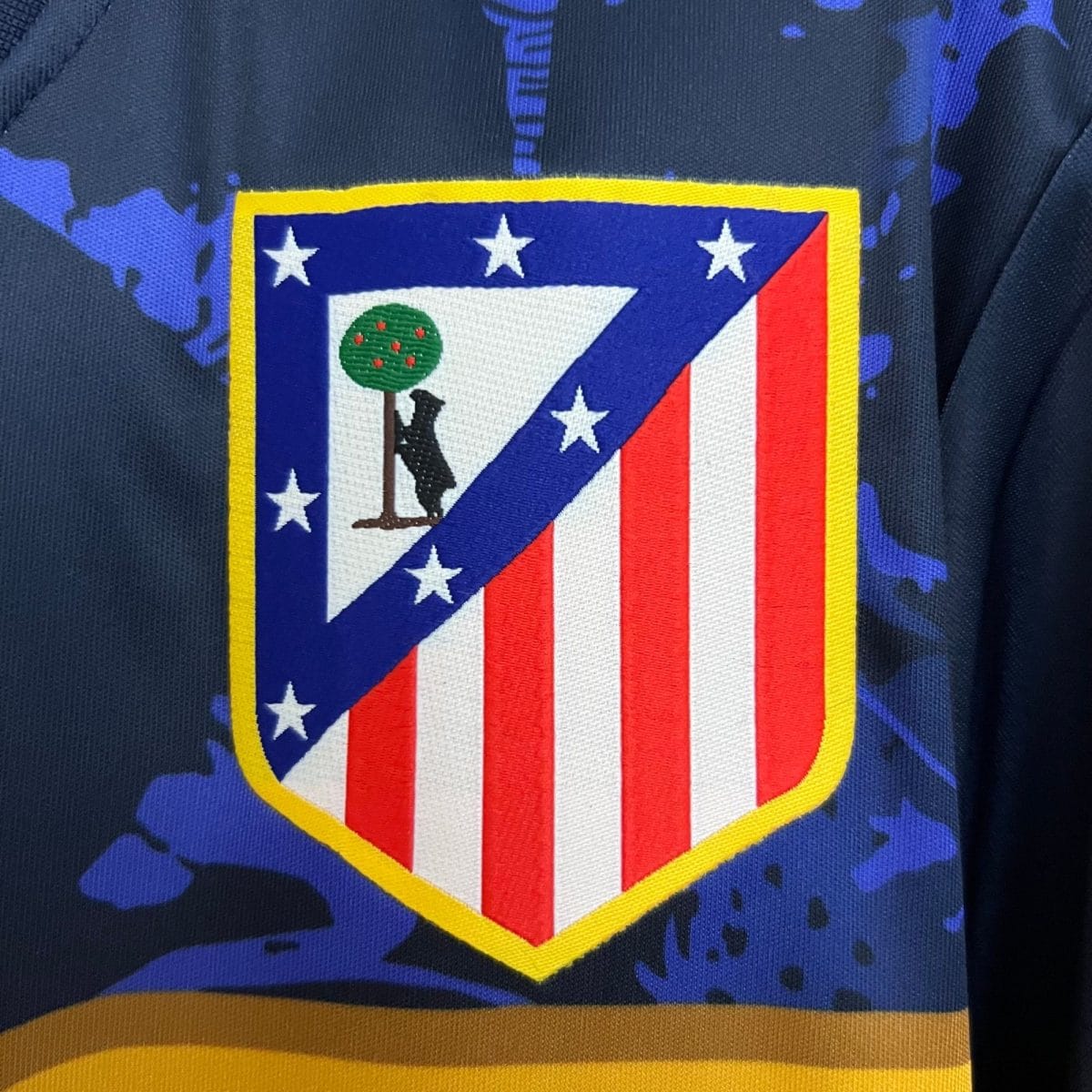 Decorative shield-shaped patch on dark fabric: blue field with white stars, red and white stripes, a diagonal blue stripe, green tree emblem, yellow border.