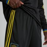 Close-up of black athletic pants with three yellow stripes along the side, a Manchester United patch near the hem, and a hand lifting the fabric.