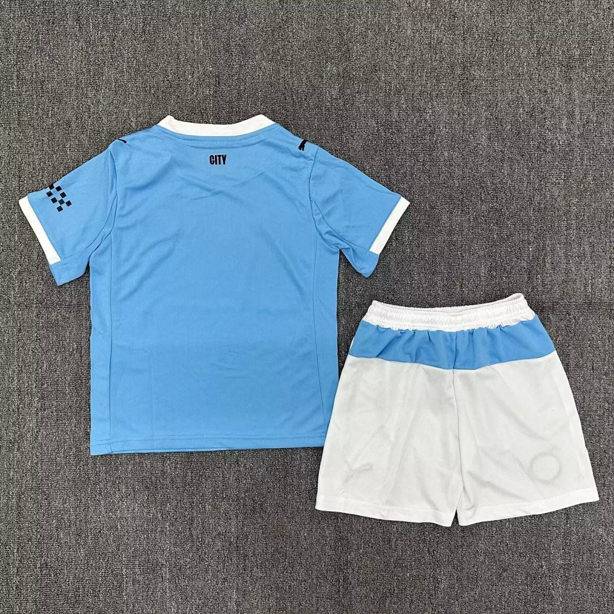 Back view of light blue soccer jersey with white collar and 'CITY' on the back, paired with matching blue and white striped shorts on a gray carpet.