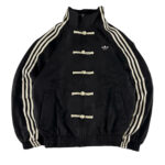 Black Adidas track jacket with cream toggle closures and three white stripes down the sleeves; small Adidas logo on the left chest.