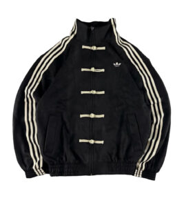 Black Adidas track jacket with cream toggle closures and three white stripes down the sleeves; small Adidas logo on the left chest.