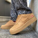 Beige suede Nike Air Force 1 sneakers with a red Supreme tag, paired with paisley-patterned pants on outdoor steps.