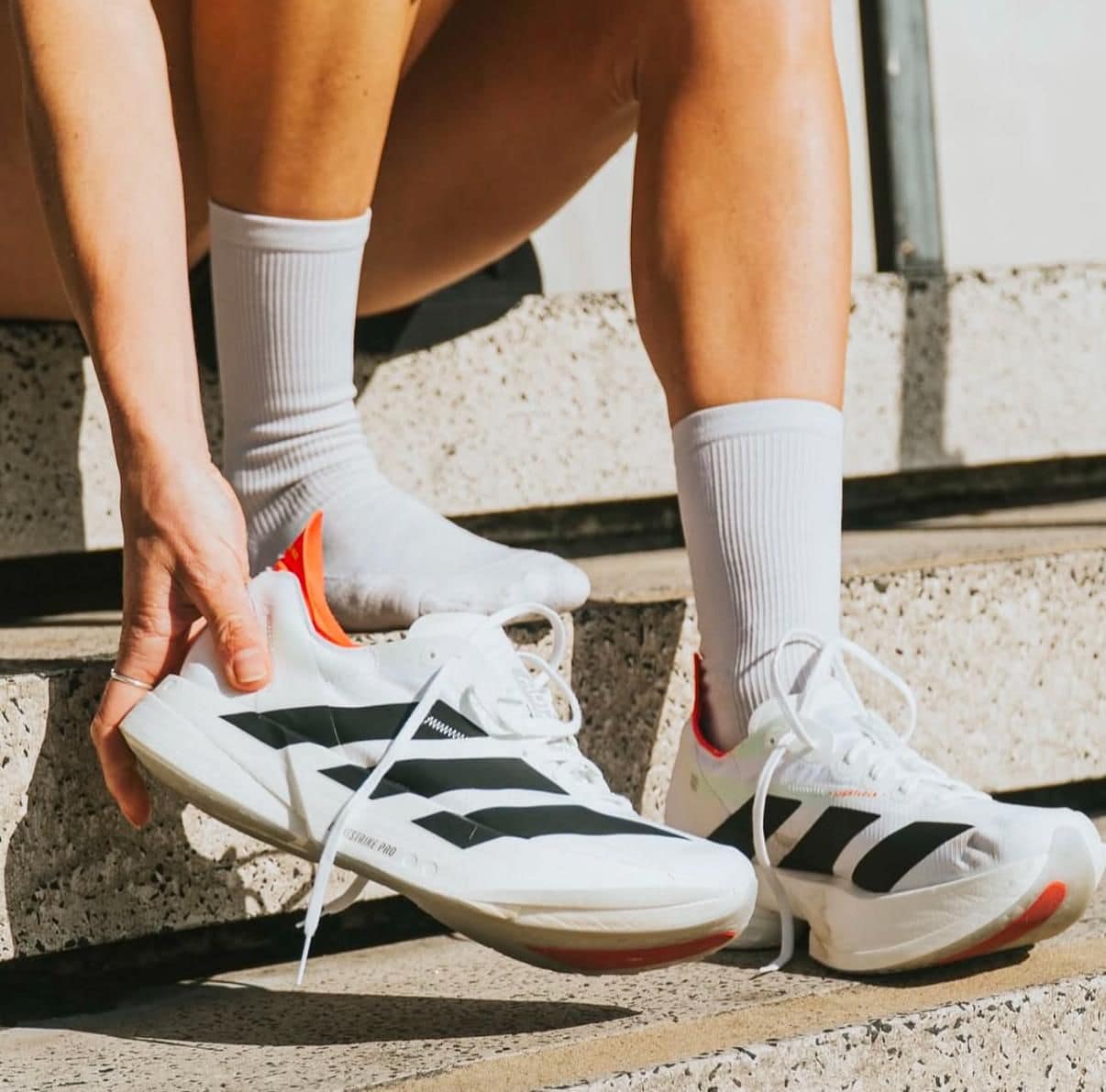 bed7e4fd-163d-417e-841a-6e08f64b7a21 | UAE SPORT STORE Person on concrete steps tying white athletic sneakers with black stripes and white socks.