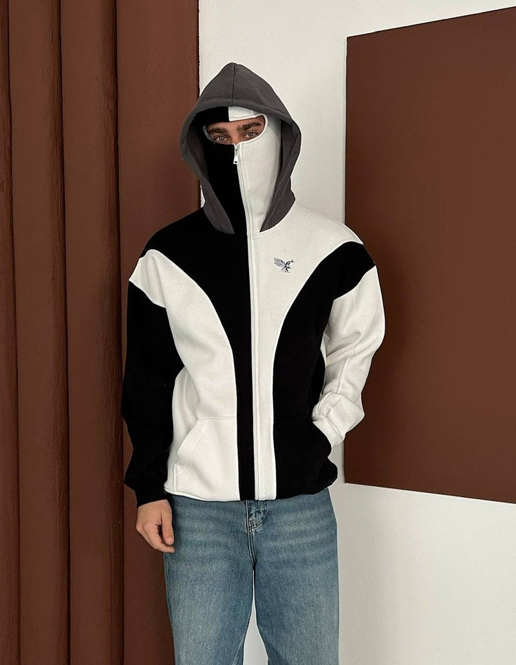 bedogiyimm_03153fd2315e9f9f75b428f02765c1dd | UAE SPORT STORE Person wearing a black and white color-block zip hoodie with a gray hood, face partially masked, standing indoors with hands in pockets.