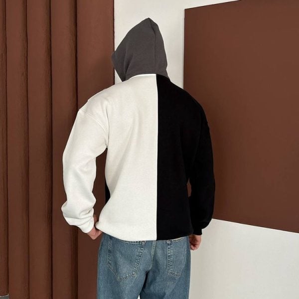 Back view of a person wearing a two-tone color-block hoodie (white left panel, black right panel) with a gray hood, paired with blue jeans, standing in a room with brown panels on the wall.