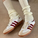 Person wearing cream ribbed socks tucked over white sneakers with red stripes and gum soles, standing on a tan carpeted floor.