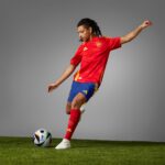 Soccer player in red Spain jersey kicking a ball on a grassy field against a gray background